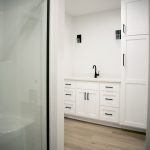 Bathroom with light gray walls, vinyl plank flooring, a glass walk-in shower, a white sink vanity, a black sink faucet, and a set of white stacked cabinets connected to the sink vanity.