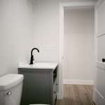 Bathroom with light gray walls, white trim, vinyl plank flooring, a white toilet, a dark gray wood sink vanity, a black sink faucet, and a white open door leading into a hallway.