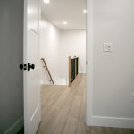 Open white door in a light gray room with vinyl plank flooring leads into a hallway with a view of the top of a staircase with light wood railings.