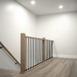 Top of staircase with light brown wood railings and black balusters, light gray walls, white trim, and vinyl plank flooring.