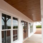 Covered front porch with a wood plank ceiling, white puck ceiling lights, and a cement porch floor attached to a home with white sliding and a brown double-door entrance with glass windows.