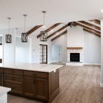 Combined kitchen and living area with a wood kitchen island, light stone countertops, vinyl plank flooring, light colored walls, a white washed brick fireplace, a black ceiling fan, and decorative wood ceiling beams.