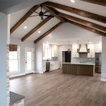 Combined kitchen and living area with a wood kitchen island, white cabinets, light blue chevron tile backsplash, light stone countertops, stainless steel appliances, vinyl plank flooring, light colored walls, a black ceiling fan, and decorative wood ceiling beams.