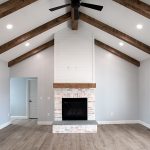 Living area with vinyl plank flooring, light colored walls, a white-washed brick fireplace with a wood beam shelf, a black ceiling fan, and decorative wood ceiling beams.