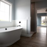 Bathroom with light blue walls, vinyl plank flooring, white trim, a white tub beside a ship lap wall, and an open door leading into a bedroom.