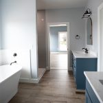 Bathroom with light blue walls, vinyl plank flooring, white trim, a white tub beside a ship lap wall, an open door leading into a bedroom, a standing shower with gray tiles, and 2 dark blue sink vanities with white counters.