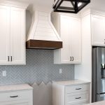 Kitchen with white cabinets, light stone countertops, a stainless steel fridge, light blue chevron tile backsplash, and a white shiplap hood range with brown wood accents.