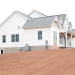 Front and left side exterior of a home with white siding, gray asphalt roofing, brick veneer, decorative shakes, a covered front porch, and a set of white basement doors.