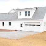 Right side exterior of a home with white siding, gray asphalt roofing, a white single-entry side door and a white garage door with windows.