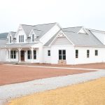 Front and right side exterior of a home with white siding, gray asphalt roofing, brick veneer, decorative shakes, a covered front porch, a white garage door with windows, and a white single-entry side door.