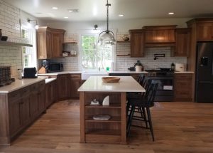 Remodeled kitchen with a brown and white island with black stool chairs, wood flooring, white tile backsplash, brown cabinets, and white marble countertops.