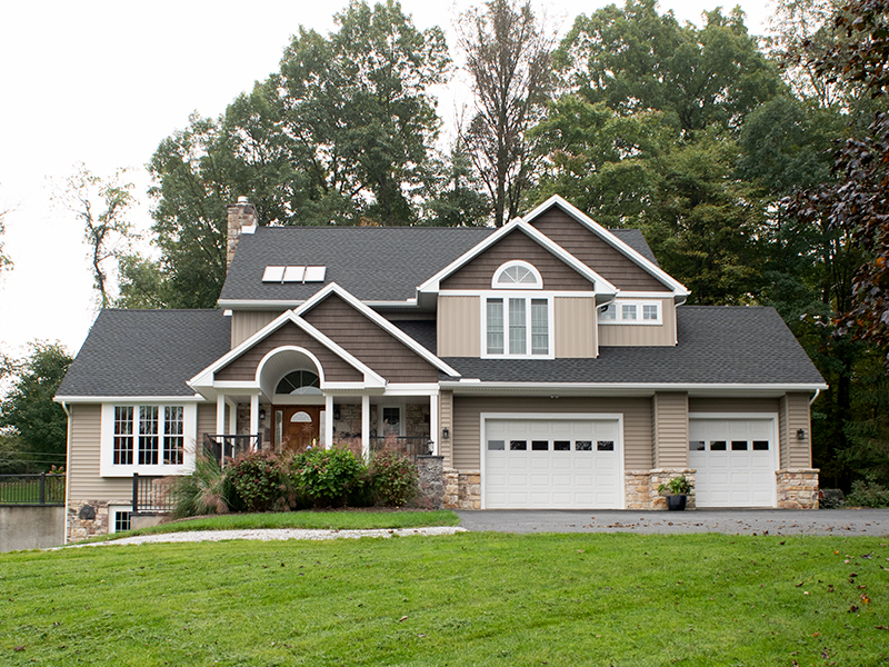 Front view of a remodeled home with gray asphalt roofing, tan vinyl siding, brown vinyl shakes, white trim, white windows, a front porch, stone accents, a stone chimney, and 2 white garage doors.