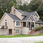 Front and left side view of a remodeled home with gray asphalt roofing, tan vinyl siding, brown vinyl shakes, white trim, white windows, a front porch with black railings, stone accents, a stone chimney, and a brown double basement door with glass panels.