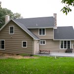 Back view of a remodeled home with gray asphalt roofing, tan vinyl siding, white trim, white windows, a stone chimney, a chimney with vinyl siding, and a stamped concrete back porch with a picnic table and outdoor loveseat.