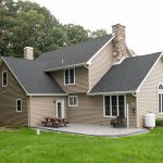 Back and left side view of a remodeled home with gray asphalt roofing, tan vinyl siding, white trim, white windows, a stone chimney, a chimney with vinyl siding, and a stamped concrete back porch with a picnic table and outdoor loveseat.