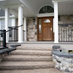 Front porch and entrance of a newly remodeled home with stone walls and a walkway leading up to the porch, black railings, white posts, a white ceiling, stone siding, and a brown wood front door with windows.