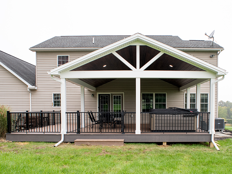 New covered deck with brown decking, white beams and posts, black aluminum railings, a black outdoor dining set, a hot tub, and a brown plank ceiling with puck lights.