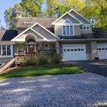 Front view of a home before its remodel with brown asphalt roofing, tan vinyl siding, white trim, white windows, a covered front porch with old wood flooring and stairs, stone accents, a stone chimney, and 2 white garage doors.