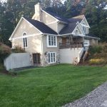 Front and left side view of a home before its remodel with brown asphalt roofing, tan vinyl siding, white trim, white windows, a covered front porch with old wood flooring and stairs, stone accents, a stone chimney, and a brown double basement door with glass panels.