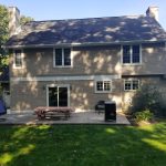 Back view of a home before its remodel with brown asphalt roofing, tan vinyl siding, white trim, white windows, a white bordered glass sliding door, a stone chimney, a chimney with vinyl siding, and a concrete back porch with a picnic table and hanging chair.