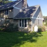 Back and left side view of a home before its remodel with brown asphalt roofing, tan vinyl siding, white trim, white windows, a stone chimney, and a white propane tank.