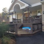 Covered front porch and entrance of a home before its remodel with an outdated and chipped wood porch painted brown, a white ceiling, stone siding, and a brown front door with windows.