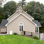 Left side view of a remodeled home with gray asphalt roofing, tan vinyl siding, white trim, white windows, a front porch with black railings, a stone chimney, a white propane tank, and black railings guarding a drop off.