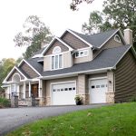 Front and left side view of a remodeled home with gray asphalt roofing, tan vinyl siding, brown vinyl shakes, white trim, white windows, a covered front porch with black railings, stone accents, stone walls lead up to the entrance, a chimney with vinyl siding, and 2 white garage doors.