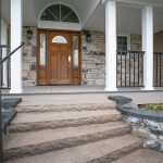 Front porch and entrance of a newly remodeled home with stone walls and a walkway leading up to the porch, black railings, white posts, a white ceiling, stone siding, and a brown wood front door with windows.