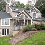 Front view of a remodeled home with gray asphalt roofing, tan vinyl siding, brown vinyl shakes, white trim, white windows, a front porch with black railings, stone accents, and a stone chimney.