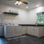 Laundry room with white walls, white cabinets, black marble countertop, a black sink, dark gray tile floor, a brown ceiling fan, and white appliances.