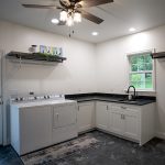 Laundry room with white walls, white cabinets, black marble countertop, a black sink, dark gray tile floor, a brown ceiling fan, and white appliances.