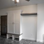 Room with white walls, dark gray tile floor, a white wardrobe with shoe cubbies underneath, a wood clothes rack, and a brown panel door.