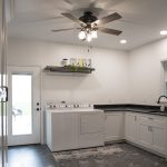 Laundry room with white walls, white cabinets, black marble countertop, a black sink, dark gray tile floor, a brown ceiling fan, white appliances, and a white door with glass.