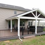 New covered deck with brown decking, white beams and posts, black aluminum railings, a black outdoor dining set, a hot tub, and a brown plank ceiling with puck lights.