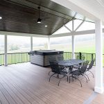 New covered deck with brown flooring, white beams and posts, black aluminum railings, a black outdoor dining set, a hot tub, and a brown plank ceiling with puck lights.