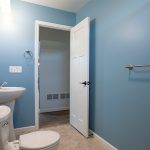 Bathroom with blue walls, a white toilet, a white sink, a white door, and tan tile flooring.