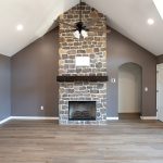 Room with brown walls, vinyl plank flooring, a white ceiling, a brown and black ceiling fan, and a brown stone electric fireplace with a floating wood shelf.