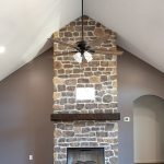 Room with brown walls, vinyl plank flooring, a white ceiling, a brown and black ceiling fan, and a brown stone electric fireplace with a floating wood shelf.