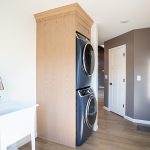 Stacked washer and dryer inside a built-in wood cabinet in a room with vinyl plank flooring, a brown accent wall, white walls, and a white sink.