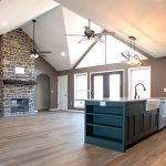 Open concept kitchen and living room with a green kitchen island that has white countertops and cabinets, vinyl plank flooring, brown walls, a white vaulted ceiling, 2 brown and black ceiling fans, and a brown stone electric fireplace with a floating wood shelf.