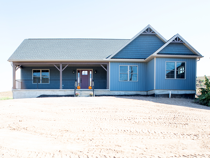 Front exterior of a new ranch-style home with blue siding, decorative blue shakes, white trim, white windows, a gray asphalt roof, and a covered front porch with black aluminum railings.