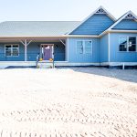 Front exterior of a new ranch-style home with blue siding, decorative blue shakes, white trim, white windows, a gray asphalt roof, and a covered front porch with black aluminum railings.