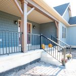 Front porch on a new, ranch-style home with concrete steps, black railings, blue siding, white windows, and brown posts.