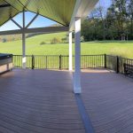 Panoramic photo overlooking a new covered deck with brown flooring, white beams and posts, black aluminum railings, a hot tub, and a brown plank ceiling with puck lights.